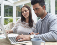 Mid adult couple paying bills at home. Mid adult woman and man are using laptop. Male and female are sitting together at table in home.